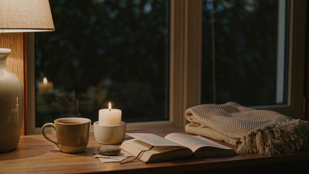 A lit candle, an open book, a ceramic mug, and a knit blanket on a nightstand by a window at night