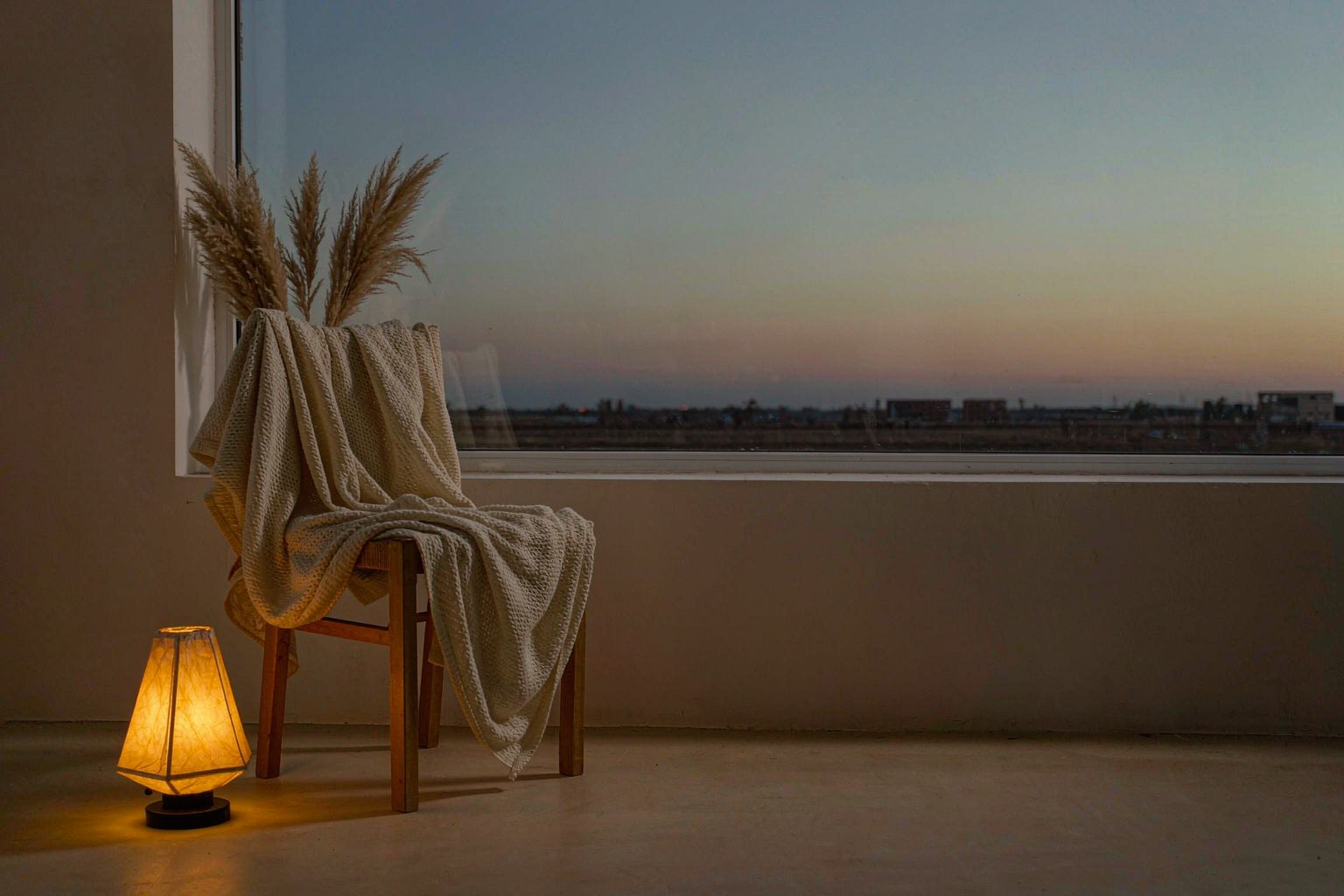 A cream knit throw over a wooden chair and a warm lamp turned low beside a dusk window.