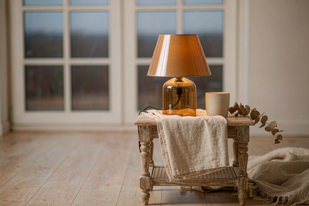 A small lamp glowing on a wooden side table beside a window at dusk, with a folded linen cloth and a cup.